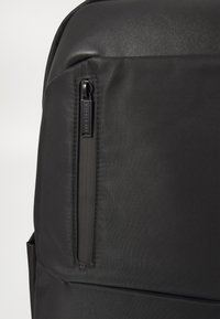 Sac à dos noir en tissu lisse et durable avec une poche zippée ; design minimaliste et lignes épurées. Couture détaillée visible.