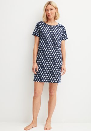 Vrouw die blootsvoets staat en een marineblauwe korte mouw jurk draagt met witte bloemenpatronen tegen een effen witte achtergrond.