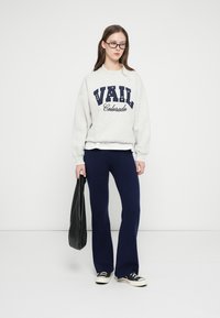 Gray sweatshirt with "VAIL Colorado" in navy lettering, paired with navy flared pants and black sneakers. Black tote bag in hand.