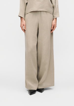 Beige Leinenhose mit weitem Bein, kombiniert mit einem passenden locker sitzenden Oberteil. Die Hose zeichnet sich durch eine entspannte Silhouette und eine glatte Textur aus, ergänzt durch spitze schwarze Schuhe.