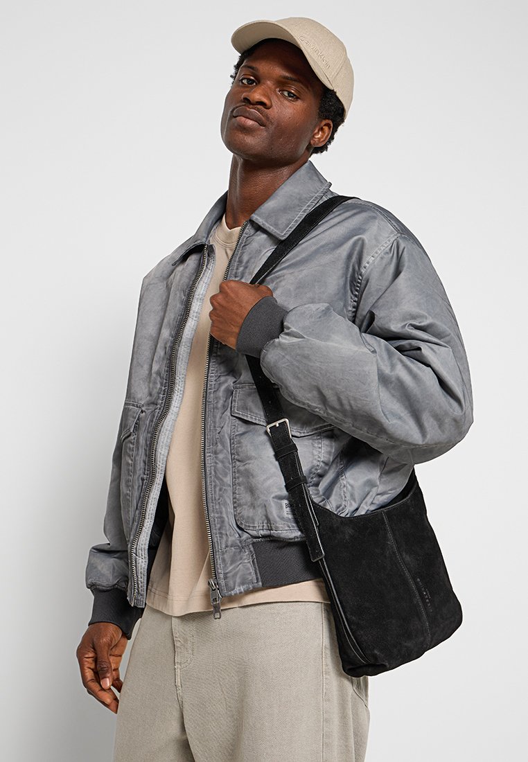 Chaqueta bomber gris con cremallera al frente y puños acanalados, combinada con un bolso de hombro de ante negro. El modelo está de pie con la mano sobre el bolso.
