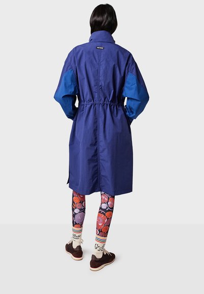 Blue long coat with elastic waist, contrasting blue sleeves, and two side pockets; paired with patterned leggings and brown shoes.