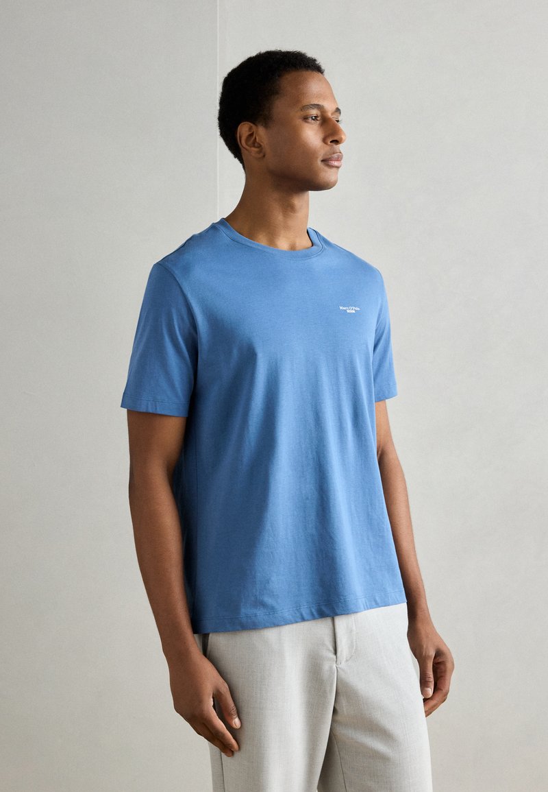 Blauwe katoenen t-shirt met een ronde hals, korte mouwen en een klein wit logo op de borst. Draag het met lichtgrijze broeken.