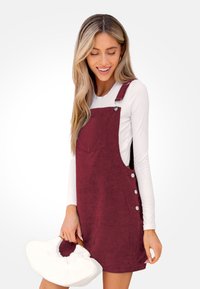 Bordeaux corduroy tuinbroek jurk met zijknopen en een voorkantzak, gecombineerd met een wit shirt met lange mouwen en een witte handtas.