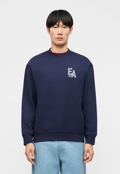 Νέος άνδρας με ίσια μαύρα μαλλιά φοράει μπλε σκούρο φούτερ με το λογότυπο "EA" στο στήθος και ανοιχτόχρωμο τζιν, μπροστά σε απλό φόντο.