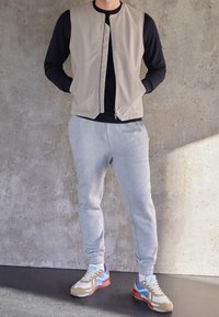 Chaleco beige claro con cremallera sobre una camisa negra de manga larga, combinado con pantalones deportivos grises y zapatillas multicolores con detalles en azul y rojo.