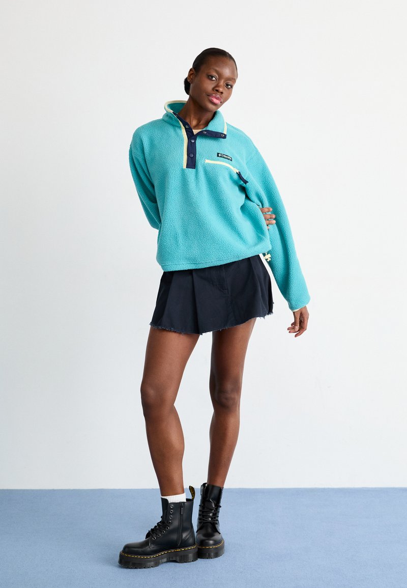 Sudadera de fleece azul claro con cuello marino y bolsillo con cremallera; combinada con una falda negra y botas negras con cordones.