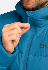 Türkis isolierte Jacke mit hohem Kragen und Reißverschluss, die ein Logo auf der Brust und einen Zugriegel zeigt, der von einer Hand gehalten wird.