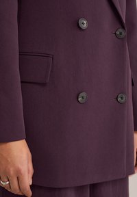 Detailní záběr na ruku vedle dvouřadého plnovínového sako v švestkové barvě se čtyřmi knoflíky a klopovou kapsou na levé straně.