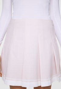 Kvinna i en ljusrosa plisserad kjol med vit fåll och långärmad vit topp, sedd från midjan till mitten av låret.