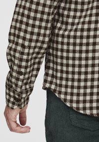 Camisa de franela a cuadros marrón y crema, con puños de botón, dobladillo curvado y etiqueta visible en el lateral. Tejido texturizado, diseño casual.