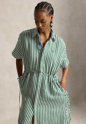 Femme aux cheveux tressés portant une robe à manches courtes rayée verticalement verte et blanche, cintrée à la taille avec des poches, regardant vers le bas.