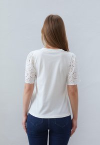 White t-shirt with floral eyelet lace puff sleeves; soft cotton fabric. Fitted design, crew neck, paired with dark blue jeans.