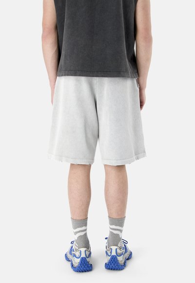 Une personne vue de dos porte un short gris clair, un débardeur gris foncé, des chaussettes grises et des baskets blanches avec des semelles bleues.