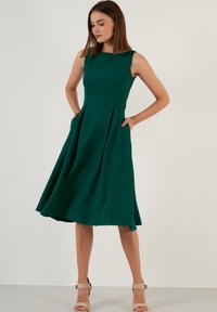 Femeie îmbrăcată într-o rochie midi fără mâneci de culoare verde pădure, stând cu mâinile în buzunare, purtând sandale cu toc până la glezne de culoare bej, pe un fundal simplu.