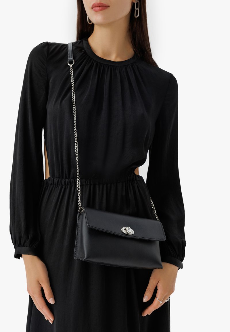 Bolso bandolera de cuero negro con herrajes plateados, tapa plegable y correa de cadena. Llevado sobre un vestido negro de manga larga.