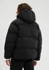 Chaqueta de plumas negra con capucha, que presenta un diseño acolchado, puños elásticos y un sutil logo en relieve en la parte posterior. Acabado suave y mate.