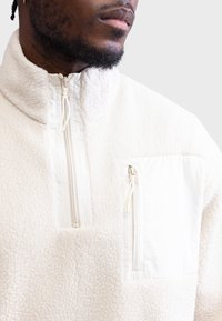 Φλις πουλόβερ σε κρεμ απόχρωση με ψηλό γιακά, μπροστινό φερμουάρ τύπου μισού και τσέπη στο στήθος με φερμουάρ που διαθέτει ομαλή αντίθετη πάνελ.