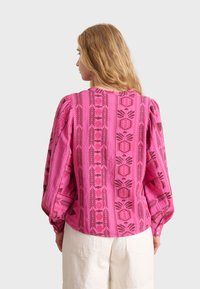 Blusa rosa a fantasia con tessuto testurizzato, maniche lunghe oversize e bottoni ai polsi. Design geometrici in rosa scuro e marrone.