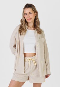 Femme aux cheveux blonds ondulés portant une chemise beige à boutons et un short assorti avec de petits imprimés de citrons jaunes, un crop top blanc en dessous.