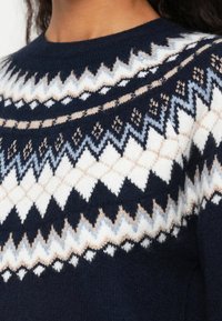 Nahaufnahme einer Person, die einen marineblauen Pullover mit einem weißen, beigen und hellblauen Zickzack- und Rautenmuster trägt.