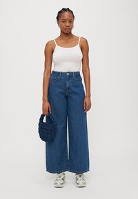 Videben bukse i blå denim med vid ben, hvit tettsittende tank topp og en navy fletteveske. Modellen har på seg mønstrede sneakers og står mot en enkel bakgrunn.
