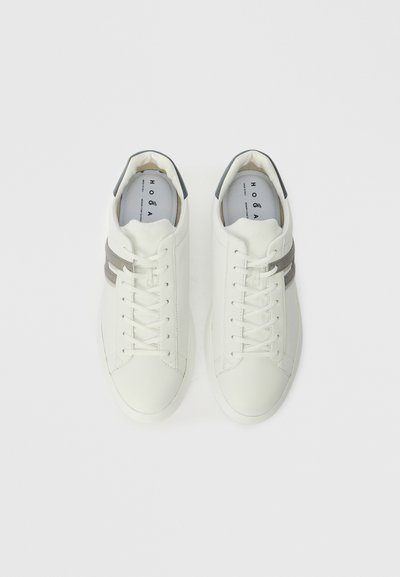 Baltas ādas sporta apavi ar pelēkiem akcentiem, plakaniem šņorēm, noapaļota purngala un minimālistisks dizains; zīmola iekšpuse ar gludu tekstūru.