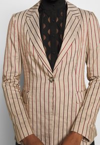 Blazer a righe in beige con strisce rosse e blu navy, chiusura a un bottone, rever a punte e due tasche anteriori. Tessuto testurizzato.