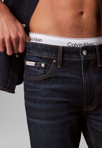 Σκούρα τζιν από ντένιμ με λογότυπο Calvin Klein, μπεζ ραφές και μπροστινή τσέπη με ορατό μεταλλικό κουμπί. Ορατή λευκή ζώνη μέσης.