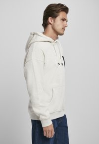 Felpa leggera color beige con cappuccio regolabile, tasca frontale e polsini a coste. Presenta una texture sottile e dettagli neri sulla tasca.
