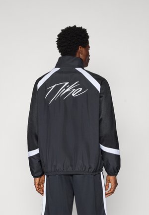 Moški v črno-beli športni jakni Nike z logotipom na hrbtu, ki stoji obrnjeno stran proti enobarvnemu sivemu ozadju.