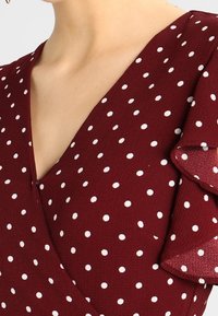 Haut en tissu bordeaux à pois blancs, avec un décolleté en V prononcé et des manches courtes à volants. Texture lisse, design décontracté.