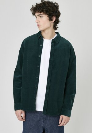 Chemise en velours côtelé vert foncé avec un col boutonné, des boutons à l'avant et des manches longues, associée à un t-shirt blanc en dessous.