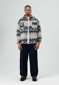 Wollen jas met geometrische patronen in zwart, beige en grijs, gecombineerd met donkere jeans met wijde pijpen en zwarte schoenen met roze veters.