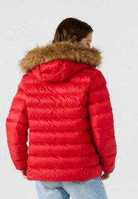 Giacca a imbottitura rossa con superficie testurizzata, trapuntatura orizzontale e bordo del cappuccio staccabile in pelliccia sintetica marrone. Vista posteriore.
