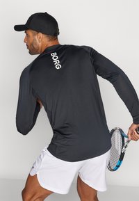 Juoda ilgomis rankovėmis sportinė maikė su užrašu "BORG" ant nugaros, derinama su baltomis šortais. Matomas teniso raketė ir aktyvi poza.