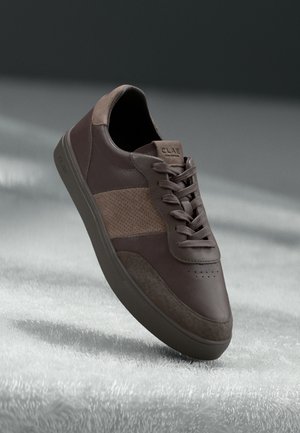 Bruine lage sneaker met suède en leren panelen, vetersluiting en een merkzool, getoond op een gestructureerd wit oppervlak met zacht licht.