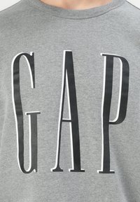Sudadera de algodón gris con letras en negrita "GAP" en negro y borde blanco. Cuello redondo clásico y acabado suave y texturizado.