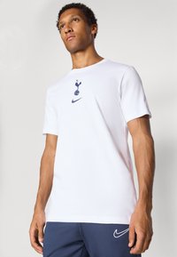 Hvid bomulds T-shirt med korte ærmer, der har et navy Spurs logo og en lille Nike swoosh på forsiden. Glat stoftekstur.