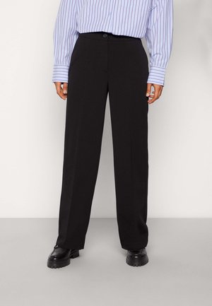 Person trägt schwarze weite Hosen, schwarze klobige Schuhe und ein langärmeliges Hemd mit lila Streifen, das in die Hose gesteckt ist, und steht vor einem einfarbigen Hintergrund.