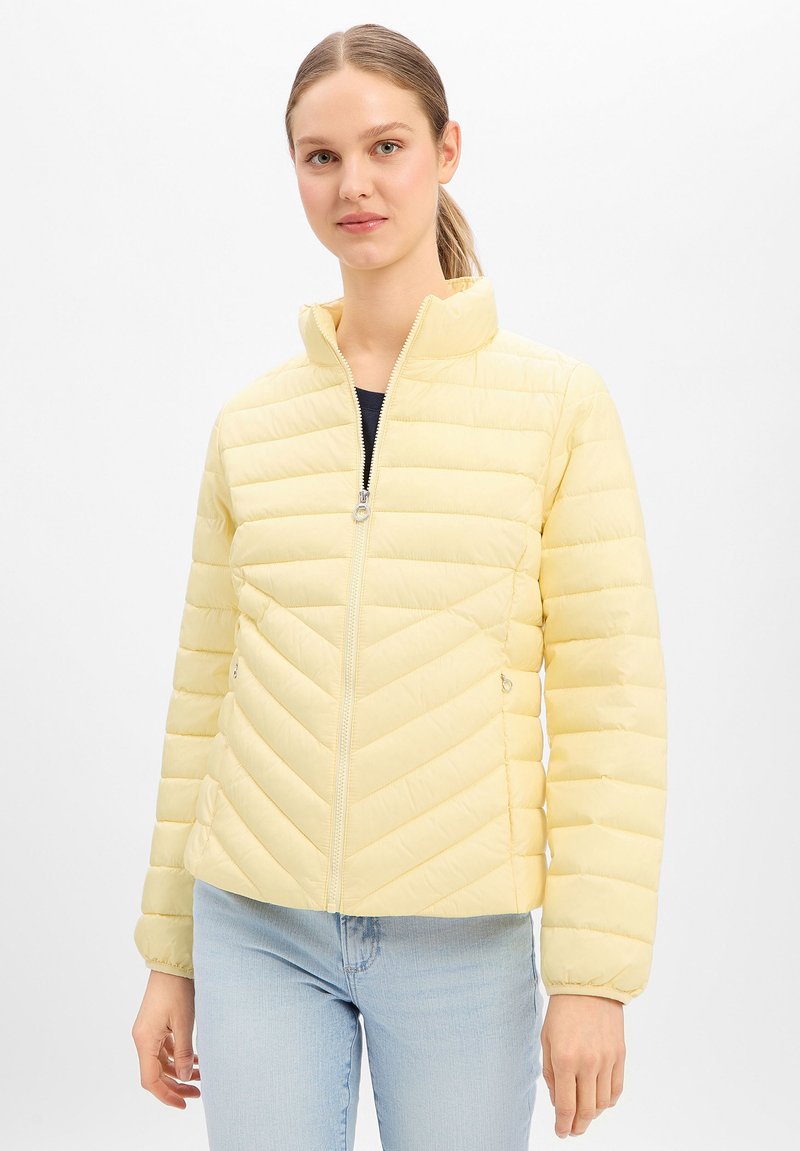 Frau mit hellbraunem Haar im Pferdeschwanz, die eine hellgelbe, gesteppte Jacke mit Reißverschluss und hellblaue Jeans vor weißem Hintergrund trägt.