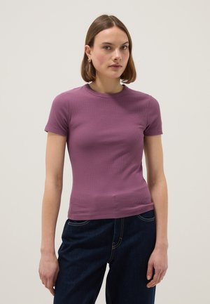 Vrouw met kort bruin haar draagt een aansluitend paars shirt met korte mouwen en donkerblauwe spijkerbroek, staand tegen een effen lichte achtergrond.