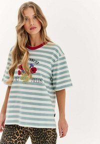 Pruhované tričko v světle modré a bílé barvě, s červeným výstřihem, květinovou grafikou s textem a krátkými rukávy. V kombinaci s kalhotami s levhartím vzorem.