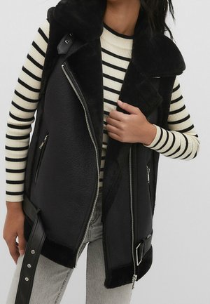 Vrouw met een zwart schapenvachtgilet met een grote kraag, gedragen over een crèmekleurige en zwarte gestreepte top met lange mouwen en lichtgrijze spijkerbroek.