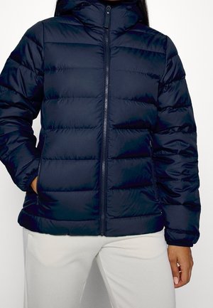 Personne portant une doudoune matelassée bleu marine avec une fermeture éclair à l'avant et un pantalon blanc, debout sur un fond blanc.