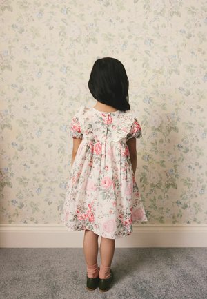 Jauna meitene ar gariem tumšiem matiem stāv pretī sienai ar ziedu tapetēm, ģērbta baltā kleitā ar rozā ziediem un volāniem.