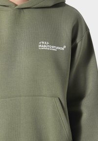 Oliivinvihreä huppari, joka on valmistettu pehmeästä puuvillasekoitteesta. Varustettu kengurutaskulla ja valkoisella painatuksessa: "IRASUTO STUDIOS" sekä englanniksi että japaniksi.