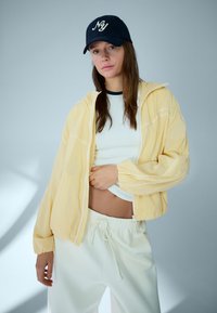 Mlada ženska z mornarsko modro kapo, rumeno jakno z zadrgo, belim kratkim topom in belimi hlačami z vrvico, stoji ob enobarvnem svetlem ozadju.