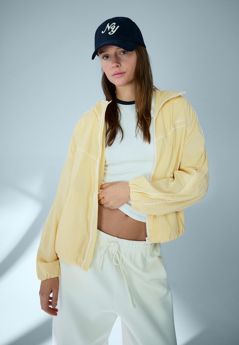 Mlada ženska z mornarsko modro kapo, rumeno jakno z zadrgo, belim kratkim topom in belimi hlačami z vrvico, stoji ob enobarvnem svetlem ozadju.