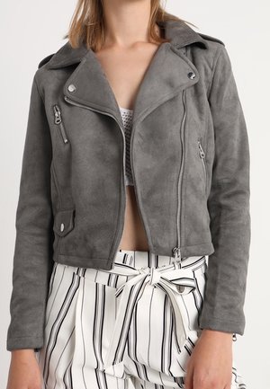 Femme portant une veste biker en daim gris avec des détails zippés et un pantalon blanc taille haute à rayures noires verticales avec un nœud de ceinture en tissu.